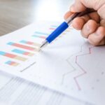 A close-up of a hand with a pen analyzing data on colorful bar and line charts on paper.