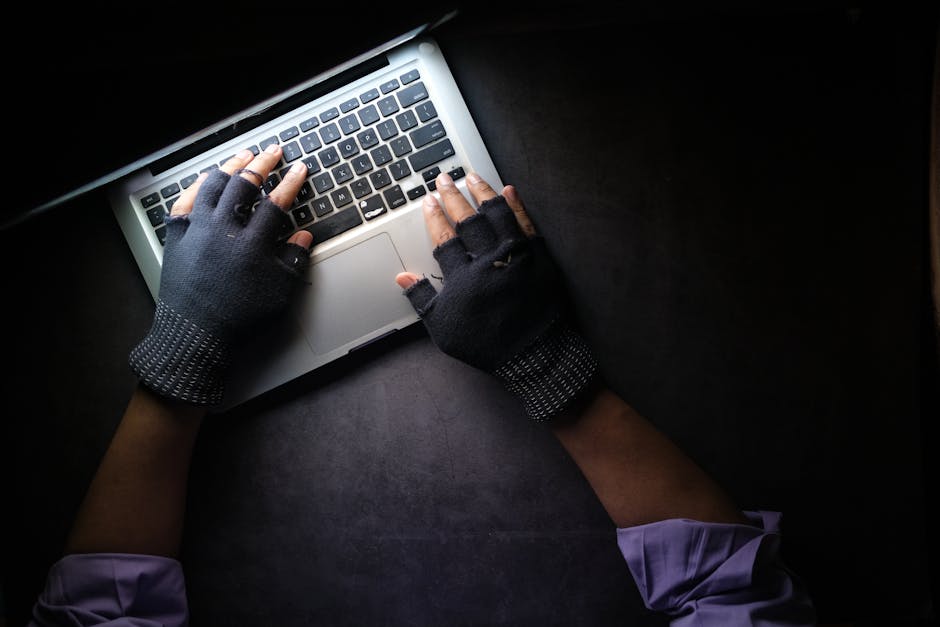 Hacker in fingerless gloves typing on laptop keyboard from above in a dark setting.